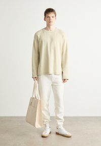 Beige hosszúujjú pulóver, fehér melegítőnadrág, semleges színű táska és fehér sportcipő barna részletekkel, egy világos, texturált padlón állva.