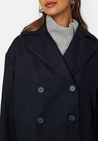 Navy modrý dvouřadý kabát s velkými knoflíky, širokými klopami a měkkou texturou. Nositý přes světle šedý žebrovaný rolák.