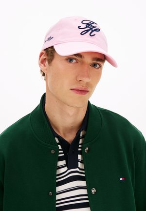 Noor mees, kes kannab roosat nokamütsi musta logoga, tumerohelist jope ja tumesinise-valge triibulise polosärgiga valge tausta ees.