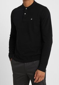 Černá polo košile s dlouhým rukávem z bavlny s dvěma knoflíky a malým vyšitým logem na levé části hrudi. Hladká textura.