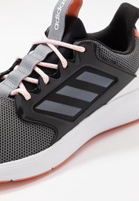 Detalle en primer plano de una zapatilla Adidas en gris y negro con cordones blancos y naranjas, parte superior de malla, logo negro de tres rayas y suela blanca con tacos naranjas.