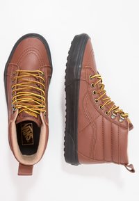 Hnědé kožené vysoké tenisky s žlutými tkaničkami, texturovaný povrch, polstrovaný límec a černá gumová podrážka. Logo Vans je viditelné uvnitř.