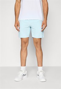 Calções desportivos azul claro feitos de um material liso e leve, combinados com ténis brancos e meias brancas, de pé contra um fundo simples.