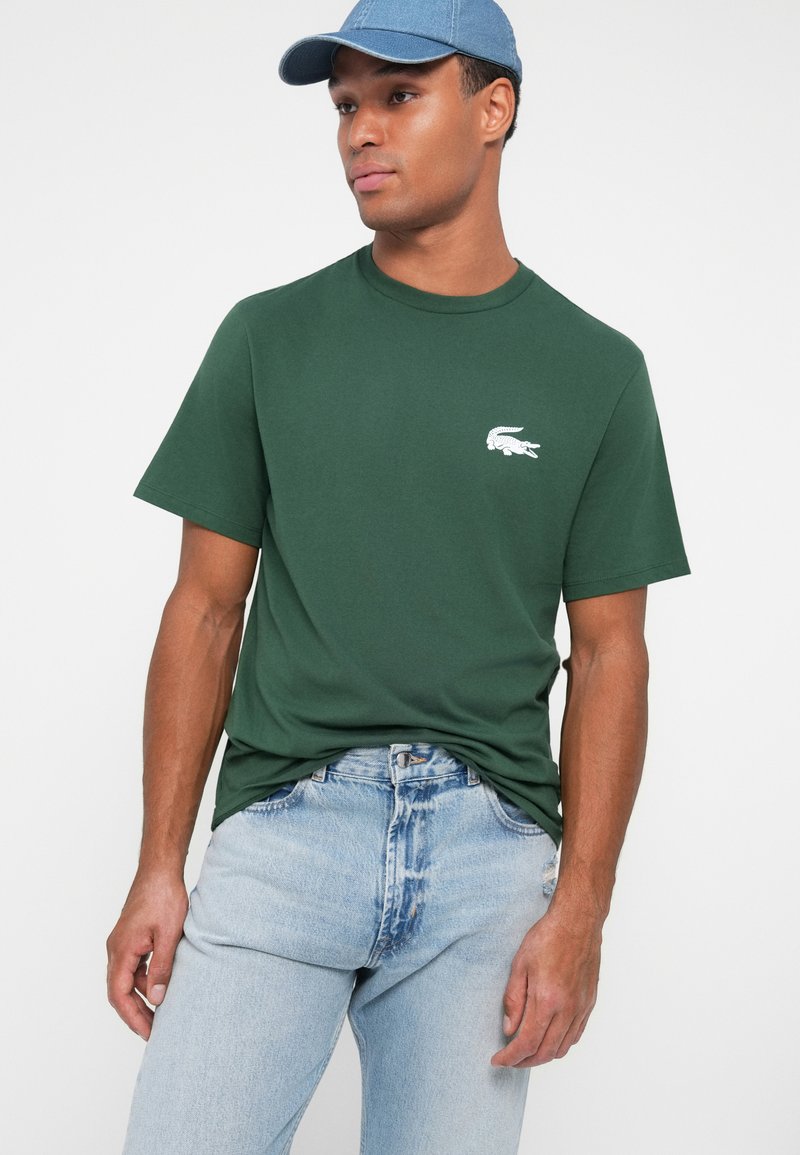 Grünes Baumwoll-T-Shirt mit kurzen Ärmeln, versehen mit einem weißen gestickten Logo auf der Brust. Getragen mit hellblauen Jeans und einer Denim-Mütze.