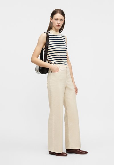 Γυναίκα που φοράει αμάνικη ριγέ μπλούζα, μπεζ παντελόνι σε φαρδιά γραμμή με ψηλή μέση, καφέ παπούτσια και μαύρη τσάντα ώμου, στέκεται με το ένα χέρι στην τσέπη.