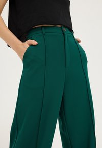 Pantalon vert taille haute en tissu lisse. Comprend des poches latérales, une fermeture à bouton et des plis marqués et repassés à l'avant.