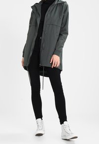 Personne portant une veste à capuche vert foncé, un pantalon noir et des baskets montantes blanches, se tenant devant un fond clair uniforme.