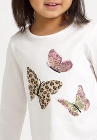 Camicetta bianca a maniche lunghe con tre design di farfalle: una con stampa leopardata, una in paillettes rosa e una in paillettes oro-verdi.