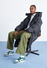 Svart puffjacka med överdimensionerad design och huva, kombinerad med gröna byxor och gröna-och-vita Nike-sneakers, sittande på en stol.