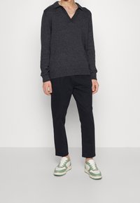 Pull en maille gris avec un col en V et des poignets côtelés, associé à un pantalon noir fuselé et des baskets blanches avec des accents verts.