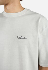 Lys grå bomulds T-shirt med rund hals; har et sort kursiv logo "Peyador" på venstre brystområde. Korte ærmer, glat tekstur.