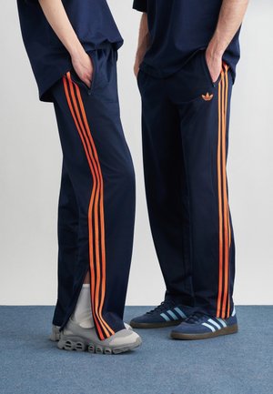 To personer står iført marineblå treningsbukser med oransje striper og lommer; den ene har på seg grå joggesko, den andre marineblå joggesko med blå striper.