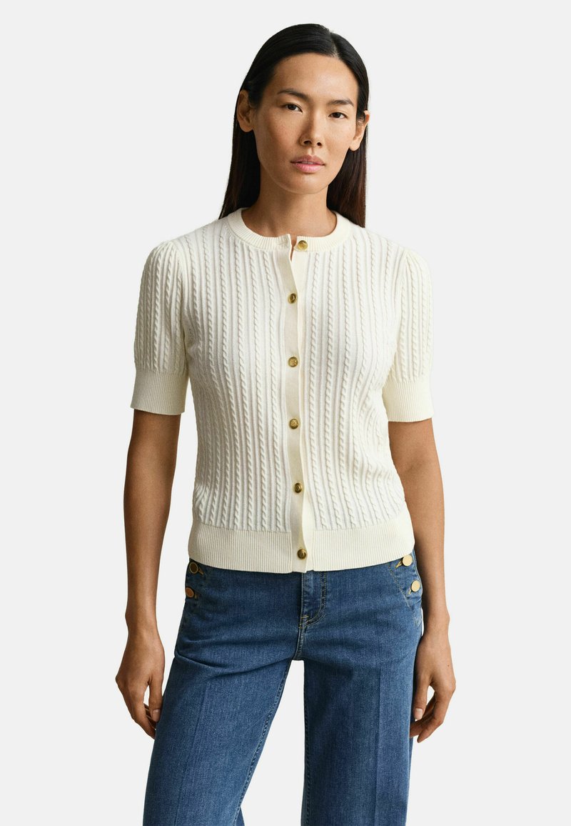 Femme portant un cardigan à manches courtes blanc en maille torsadée avec des boutons dorés et un jean bleu taille haute avec des détails de boutons.