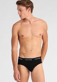 Schwarze Baumwollslips mit einem weichen Bund, der mit weißen "Calvin Klein"-Markenaufdruck versehen ist, eng anliegende Passform und glatte Textur. Modell schaut nach rechts und trägt kein Shirt.
