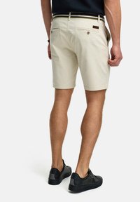 Beige op maat gemaakte shorts met een platte voorkant, twee achterzakken met knopen en een contrasterende gestreepte riem. Gemedieerd met zwarte sneakers.