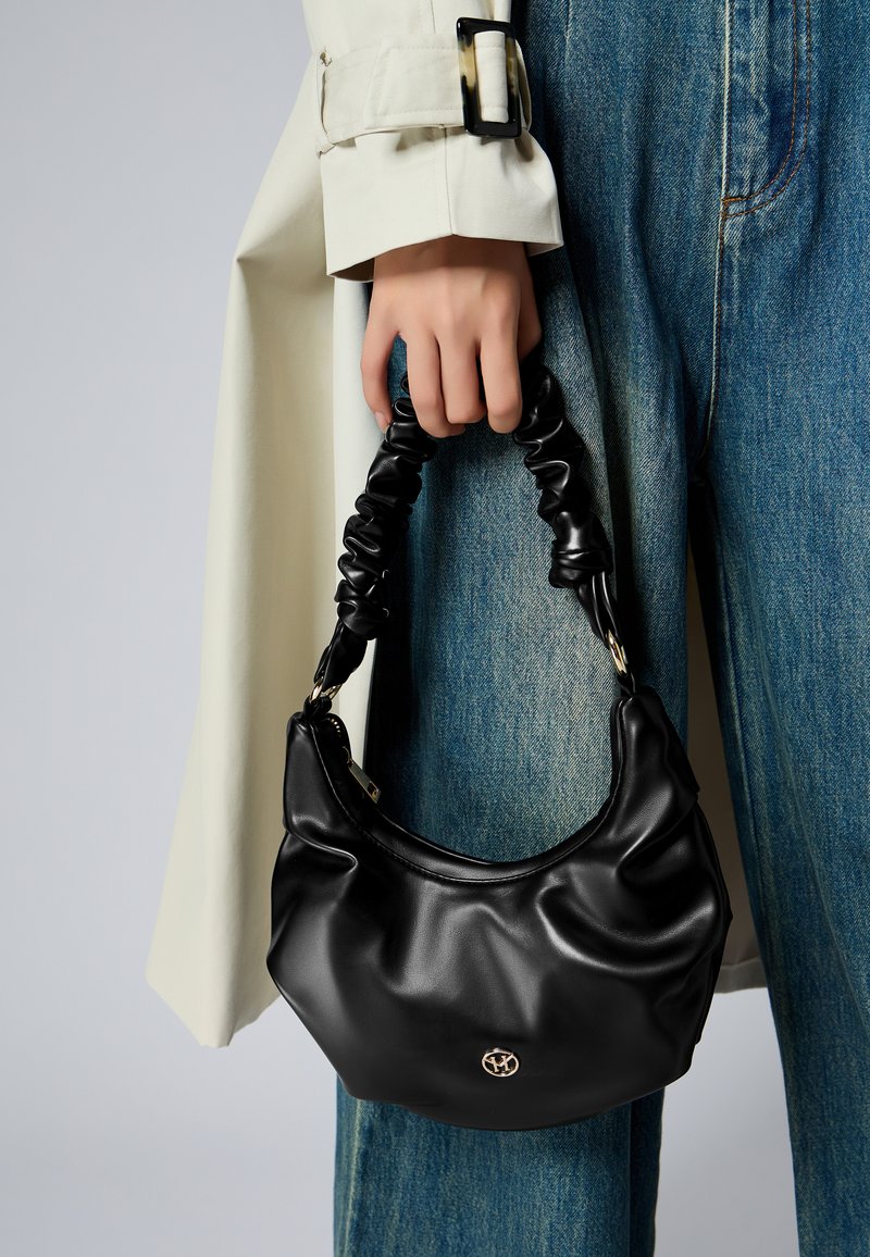 Sac à main en cuir noir avec une poignée froncée, présentant une texture lisse et un petit accent logo doré à l'avant.