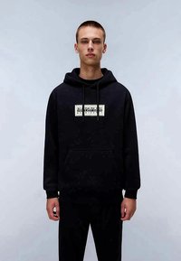 Sweatshirt preto com capuz e um bolso frontal. Apresenta um patch de logotipo creme no centro. O tecido parece suave e texturizado. Ajuste casual.
