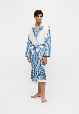 Ein Mann steht barfuß in einem weißen Bademantel mit blauen Paisley-Mustern, der an der Taille gebunden ist, vor einem einfarbigen hellen Hintergrund.