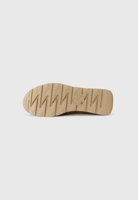 Semelle de chaussure en caoutchouc beige avec un motif de crampons en zigzag et un logo en relief. La texture lisse et la forme arrondie définissent le design.
