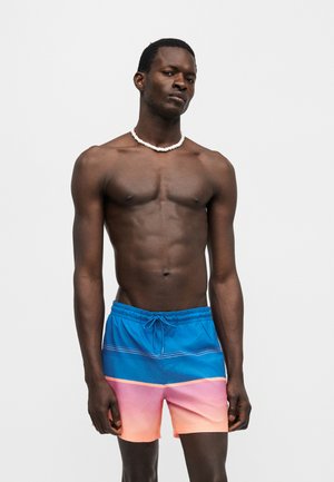 Homme torse nu portant un short de bain dégradé bleu et rose et un collier de coquillages blanc, debout devant un fond clair uni.