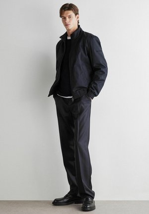 Jeune homme debout, les mains dans les poches, portant une veste noire zippée, un pantalon noir et des chaussures noires, devant un mur clair uni.