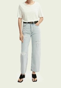 Donna che indossa una camicia bianca a maniche corte, jeans strappati azzurri a vita alta con una cintura nera e sandali neri con tacco.