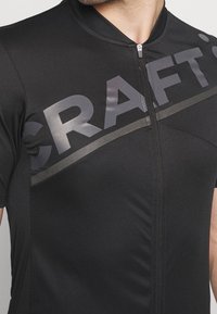 T-shirt de sport noir à manches courtes, avec une fermeture éclair et un logo "CRAFT" brillant sur la poitrine. Fabriqué dans un matériau lisse et ajusté.