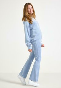 Ung gravid kvinna som ler, iklädd ljusblå tröja och byxor, poserar från sidan med vita sneakers framför en enfärgad bakgrund.