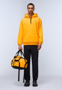 Felpa gialla con cappuccio e tasca a marsupio, con lacci neri regolabili, abbinata a una borsa a tracolla nera e gialla, pantaloni neri e scarpe.