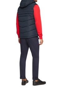 Dunkelblauer Puffer-Westen mit Kapuze, getragen über einem roten Sweatshirt. Dunkelblaue Hose und schwarze Sneaker vervollständigen das Outfit. Texturierte Nähte sind sichtbar.
