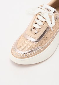 Lys beige sneakers med metalliske detaljer, vevd tekstur og hvite snørebånd. Har en tykk hvit såle og kontrasterende sømmer.