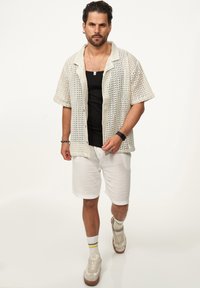 Leichtes, offen gestricktes beiges T-Shirt mit kurzen Ärmeln, schwarzes Tanktop, weiße Shorts und beige Sneakers mit gelb gestreiften Socken.