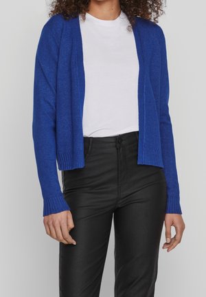 Mujer con un cárdigan azul real, camiseta blanca y pantalones negros de cintura alta, posando frente a un fondo claro y liso.