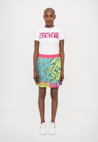 T-shirt bianca con testo in rosa brillante "VERSACE JEANS COUTURE"; gonna multicolore con un mix di motivi blu, verdi e rosa. Sneakers bianche.