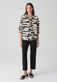 Camicia bianca e nera con motivi astratti, davanti con bottoni, maniche lunghe; abbinata a pantaloni neri e sandali neri con cinturini.