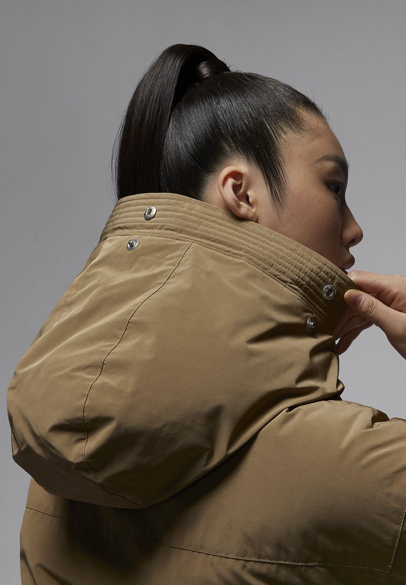 Mujer con una coleta alta ajustando el cuello de una chaqueta marrón claro con botones a presión metálicos frente a un fondo gris liso.