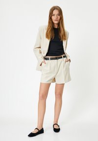 Beige bleiser muotoilultaan, musta tiukka toppi, beige laskostetut shortsit ja mustat tasapohjaiset kengät. Sileät kankaat; klassinen leikkaus modernilla siluetilla.