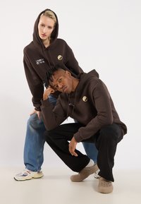 Two models wear brown hoodies featuring a yin-yang emblem and white text. One model has blue jeans, the other black pants, with varied footwear.