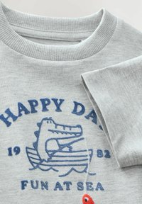 Grauer Sweatshirt mit geripptem Kragen, das ein blaues gesticktes Design eines Krokodils in einem Boot und einen roten Fischakzent zeigt.