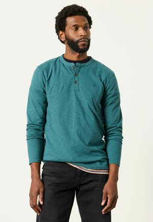 Mann mit Bart und kurzen lockigen Haaren trägt ein petrolfarbenes langärmeliges Henley-Shirt und schwarze Hosen, blickt nach rechts vor einfachem Hintergrund.