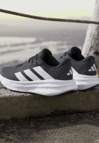 Paire de chaussures de course Adidas noires et blanches avec des semelles blanches épaisses posée sur un rebord en pierre à l'extérieur près d’un arbre et d’une barrière en corde.