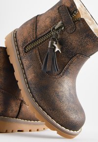 Bottes basses en similicuir marron avec une finition texturée, fermeture éclair latérale, et ornements décoratifs en forme de pompon et d'étoile. Semelle en caoutchouc avec rainures antidérapantes.