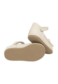 Zapatos de niño color beige con parte superior de cuero suave, cinta de velcro y un lazo decorativo; suela de goma beige texturizada con un patrón de rejilla.