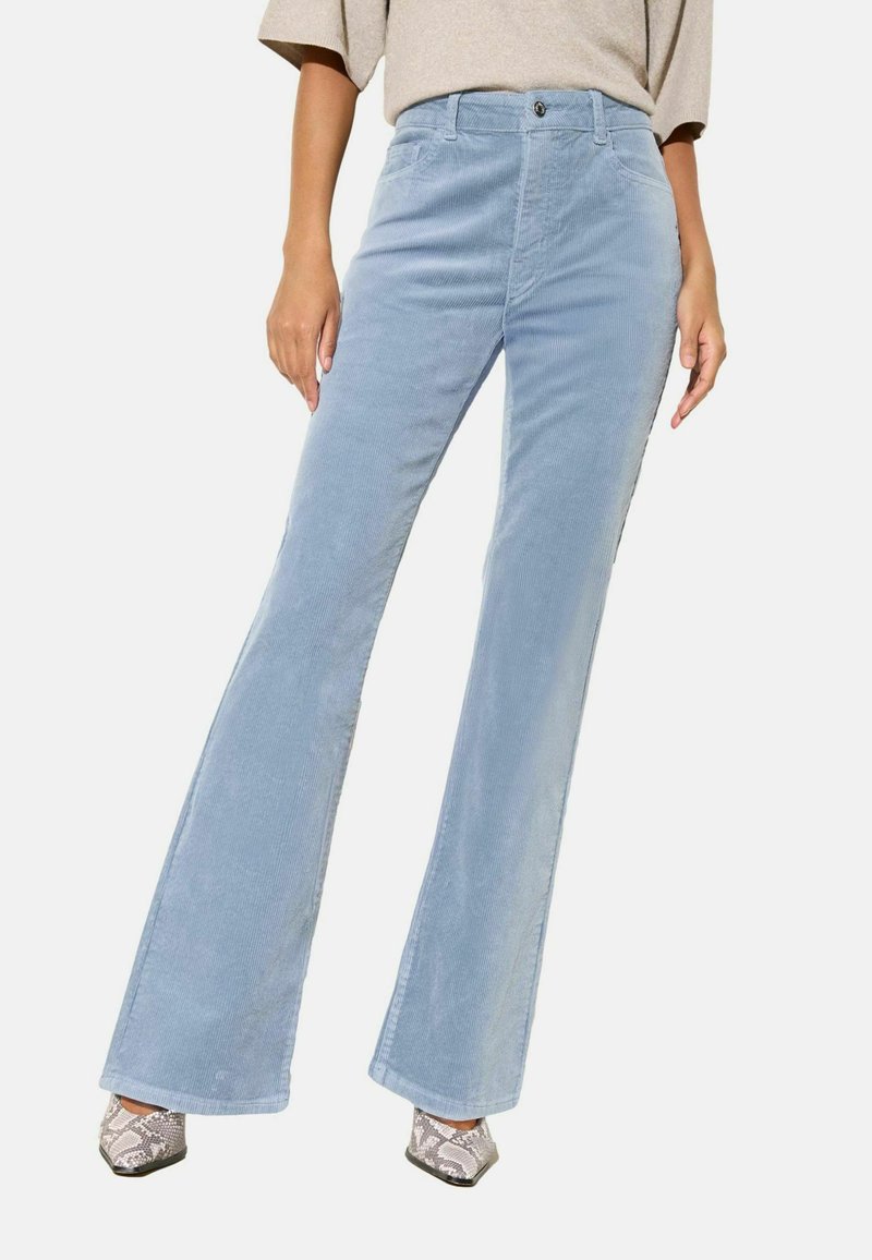 Pantalon évasé en velours côtelé bleu clair avec une fermeture boutonnée à l'avant, des poches latérales et une texture douce. Porté avec des chaussures à talons hauts à motifs.