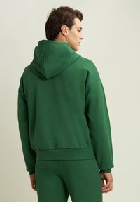 Sudadera verde hecha de tela suave y lisa, con capucha ajustable, puños acanalados y un corte relajado. Diseño de espalda sin costuras notable.