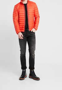 Veste matelassée rouge avec un col haut, portée sur une chemise noire, associée à un jean foncé et des bottes en cuir bleu marine à lacets avec des accents orange.