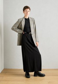 Geweven geruite blazer in beige en zwart, zwarte aansluitende top, lange zwarte rok, massieve zwarte schoenen, staand op een houten vloer tegen een grijze muur.