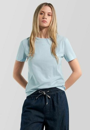 Blonde Frau trägt ein hellblaues kurzärmeliges Shirt und dunkelblaue Hose mit Kordelzug, steht mit den Händen hinter dem Rücken.