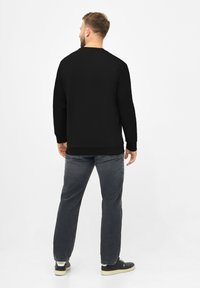 Sudadera negra de manga larga, con puños y dobladillo acanalados. Combinada con jeans grises y zapatillas negras, vista desde atrás. Fondo sencillo.