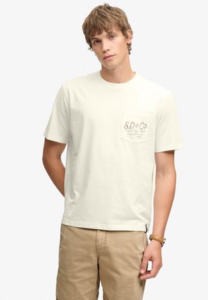 Junger Mann mit kurzen braunen Haaren, der ein weißes kurzärmeliges T-Shirt mit Brusttasche und beigen Hosen vor weißem Hintergrund trägt.